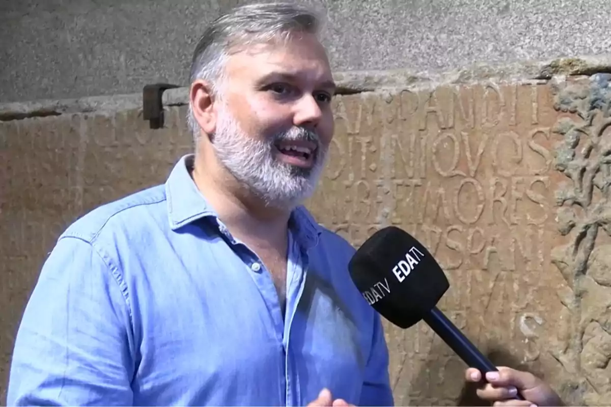 Hombre de barba canosa y camisa azul siendo entrevistado con un micrófono de EDATV frente a una pared de piedra con inscripciones antiguas Hombre de barba canosa y camisa azul siendo entrevistado con un micrófono de EDATV frente a una pared de piedra con inscripciones antiguas