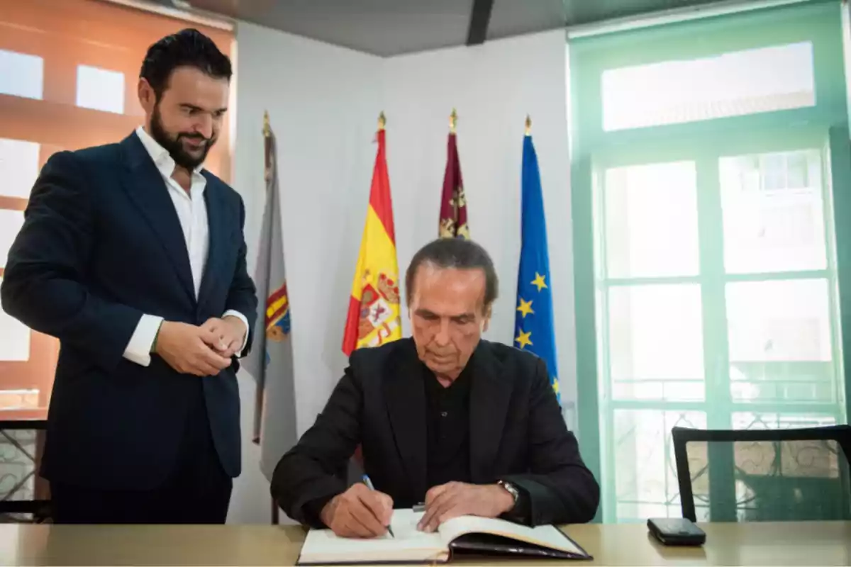 Dos hombres con traje en una oficina, uno de ellos está firmando un libro sobre una mesa y detrás hay varias banderas oficiales Dos hombres con traje en una oficina, uno de ellos está firmando un libro sobre una mesa y detrás hay varias banderas oficiales