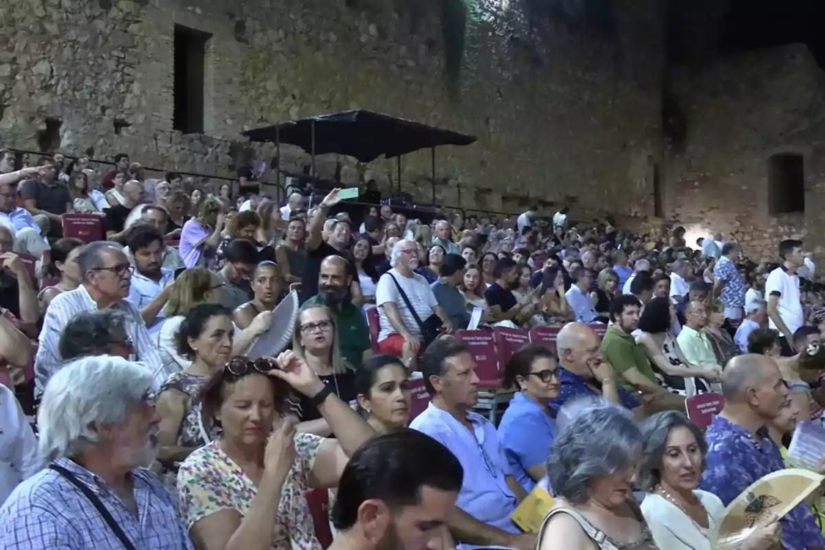 Público sentado en un auditorio al aire libre con paredes de piedra, algunas personas usan abanicos y otras conversan mientras esperan el inicio de un evento. Público sentado en un auditorio al aire libre con paredes de piedra, algunas personas usan abanicos y otras conversan mientras esperan el inicio de un evento.
