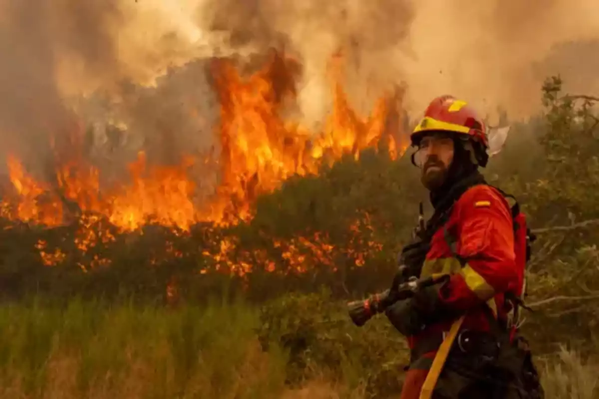 Bombero con uniforme rojo y casco observando un incendio forestal con grandes llamas y humo en el fondo