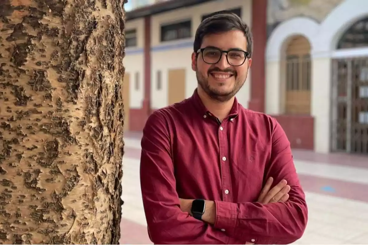 Hombre joven con gafas y camisa roja sonríe con los brazos cruzados junto a un árbol en un entorno urbano