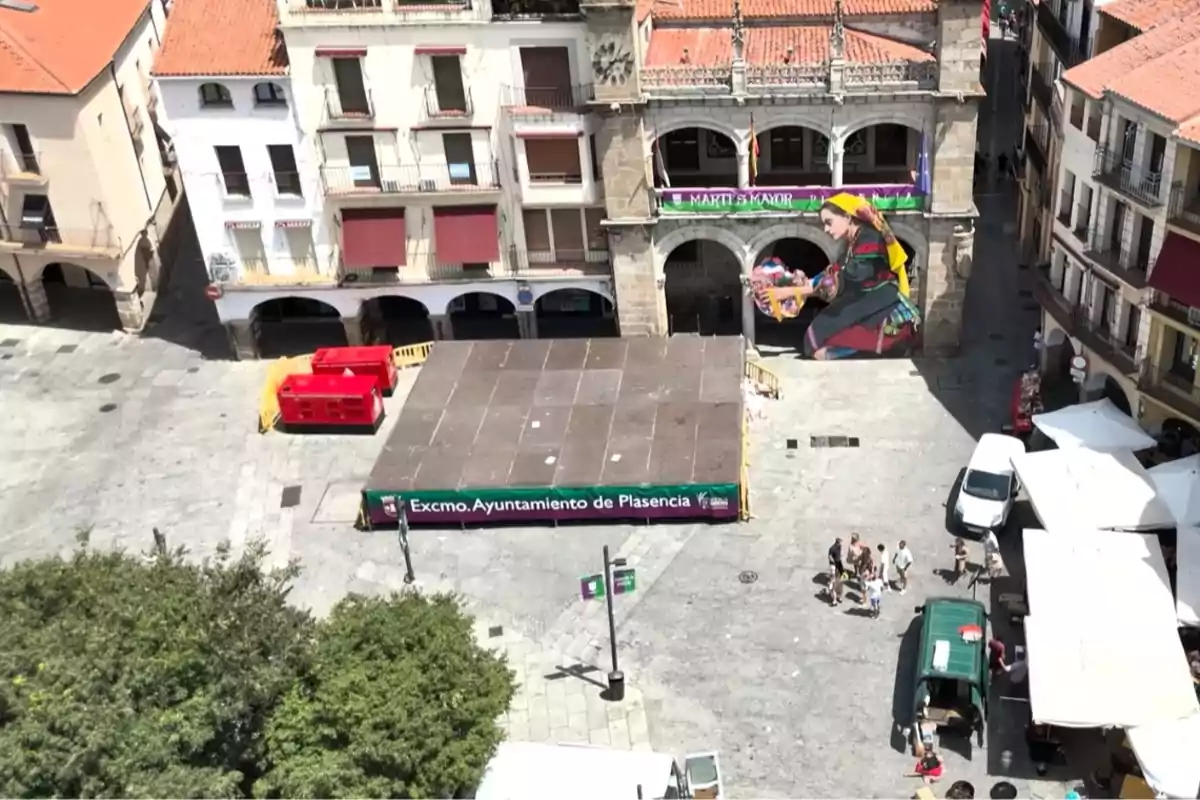 Vista aérea de una plaza con una tarima en el centro, un mural colorido de una mujer en la pared del fondo, varios edificios antiguos alrededor y algunas personas caminando cerca de puestos y vehículos. Vista aérea de una plaza con una tarima en el centro, un mural colorido de una mujer en la pared del fondo, varios edificios antiguos alrededor y algunas personas caminando cerca de puestos y vehículos.