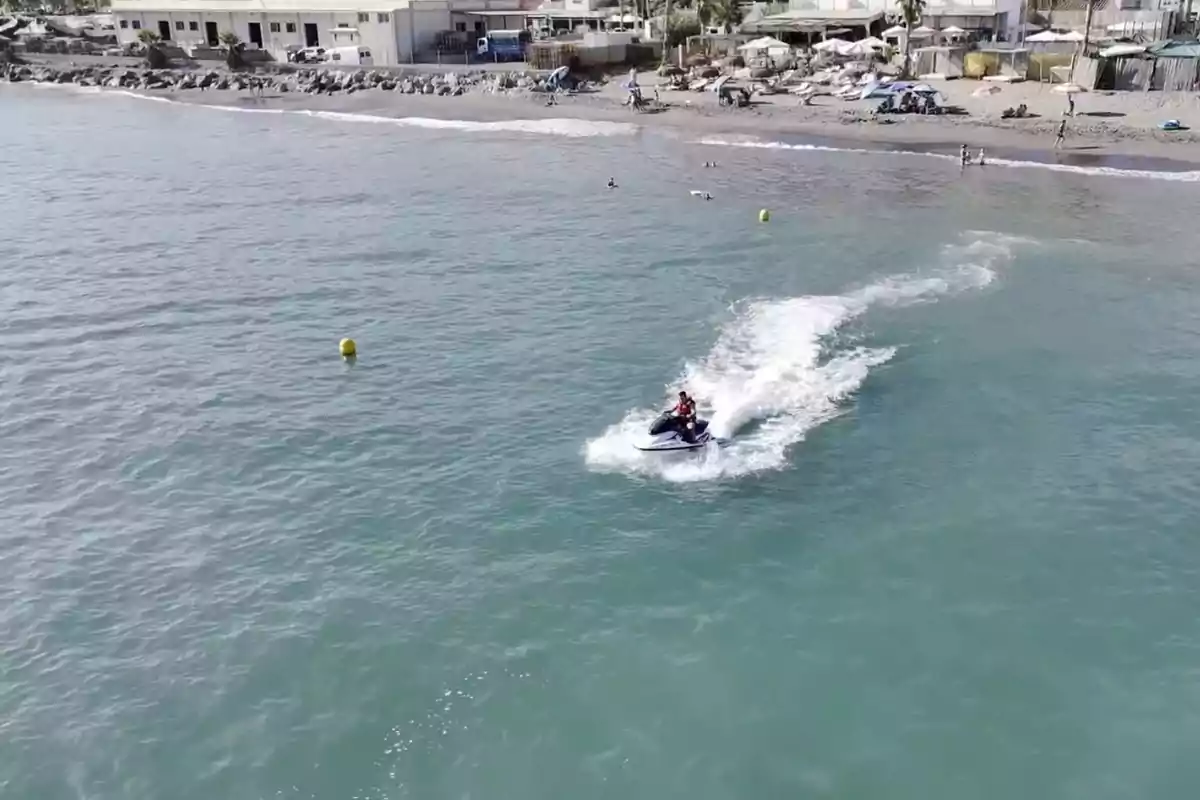 Persona conduciendo una moto acuática en el mar cerca de la orilla con algunas personas en la playa y casas al fondo Persona conduciendo una moto acuática en el mar cerca de la orilla con algunas personas en la playa y casas al fondo