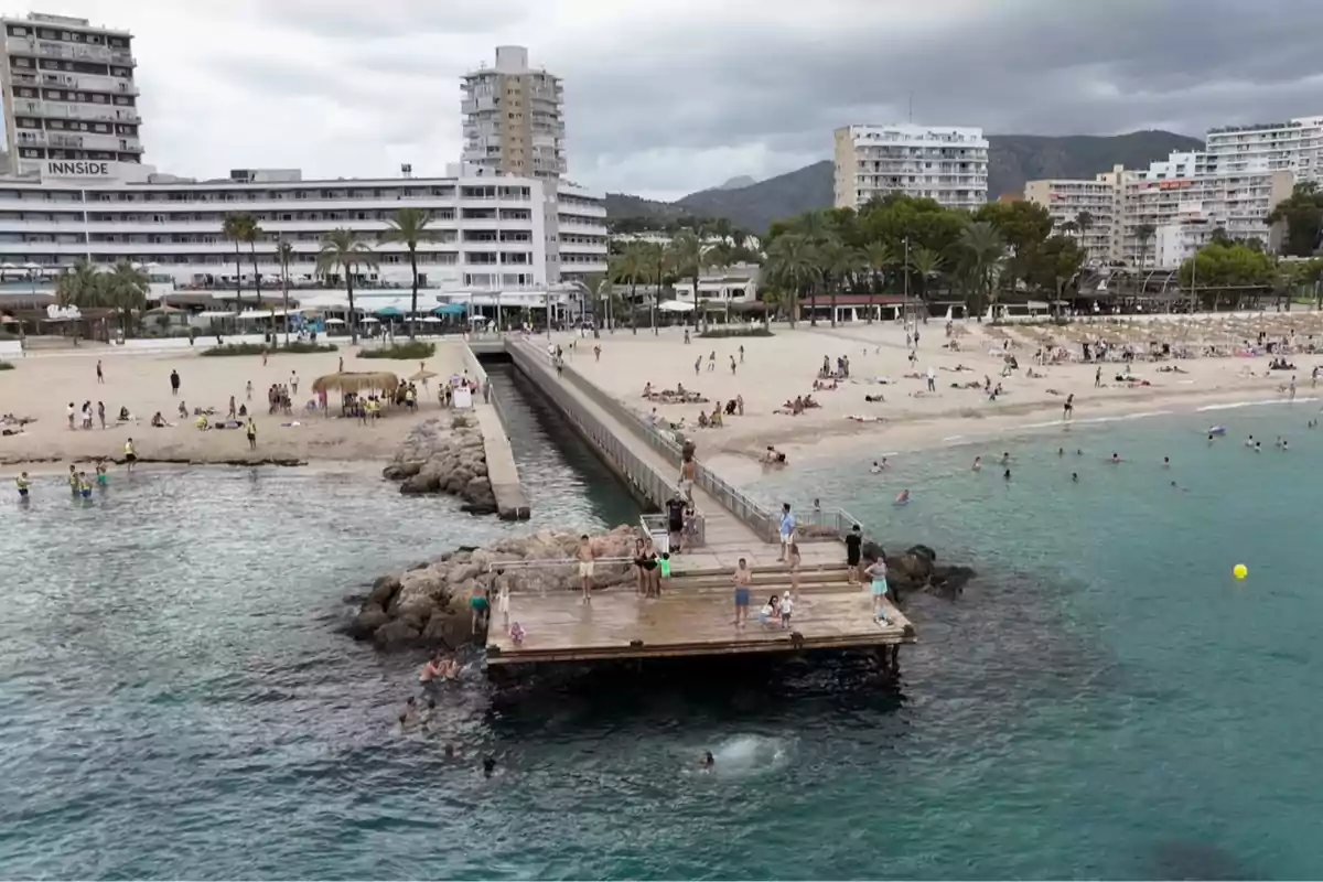 Personas disfrutando en una playa con un muelle de madera que se adentra en el mar, edificios altos y palmeras al fondo bajo un cielo nublado Personas disfrutando en una playa con un muelle de madera que se adentra en el mar, edificios altos y palmeras al fondo bajo un cielo nublado