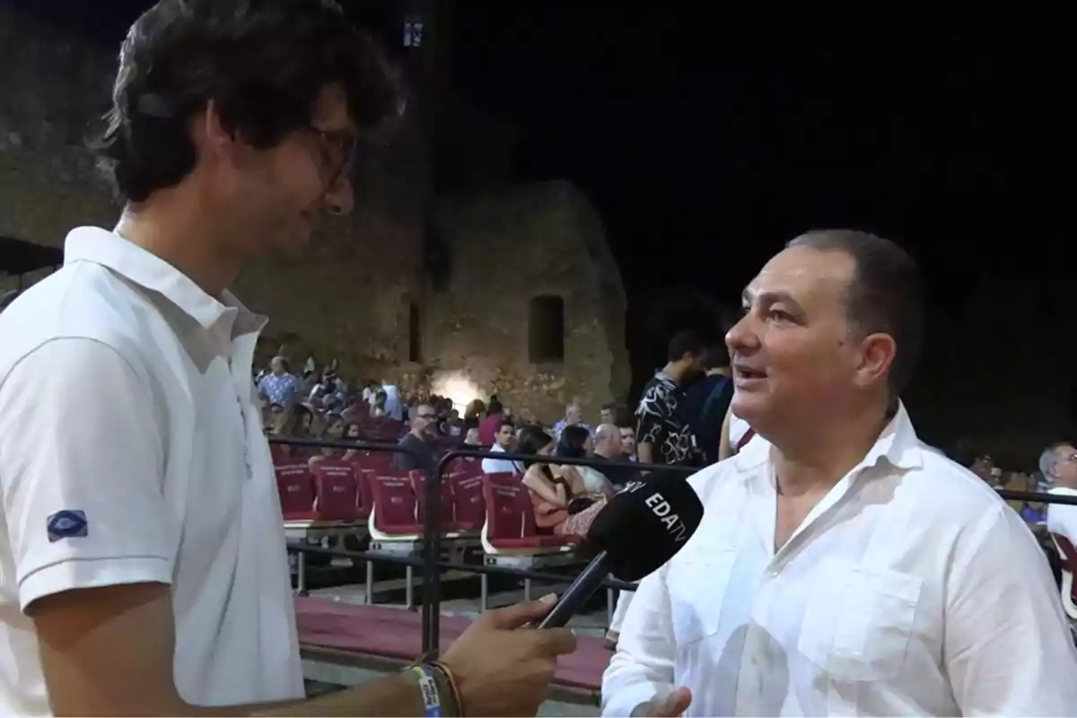Un hombre con micrófono entrevista a otro hombre en un recinto al aire libre con público sentado al fondo Un hombre con micrófono entrevista a otro hombre en un recinto al aire libre con público sentado al fondo