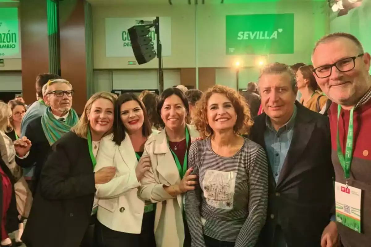 Un grupo de personas sonríe y posa para una foto en un evento en un recinto con carteles verdes en las paredes que dicen Sevilla y Corazón del cambio Un grupo de personas sonríe y posa para una foto en un evento en un recinto con carteles verdes en las paredes que dicen Sevilla y Corazón del cambio