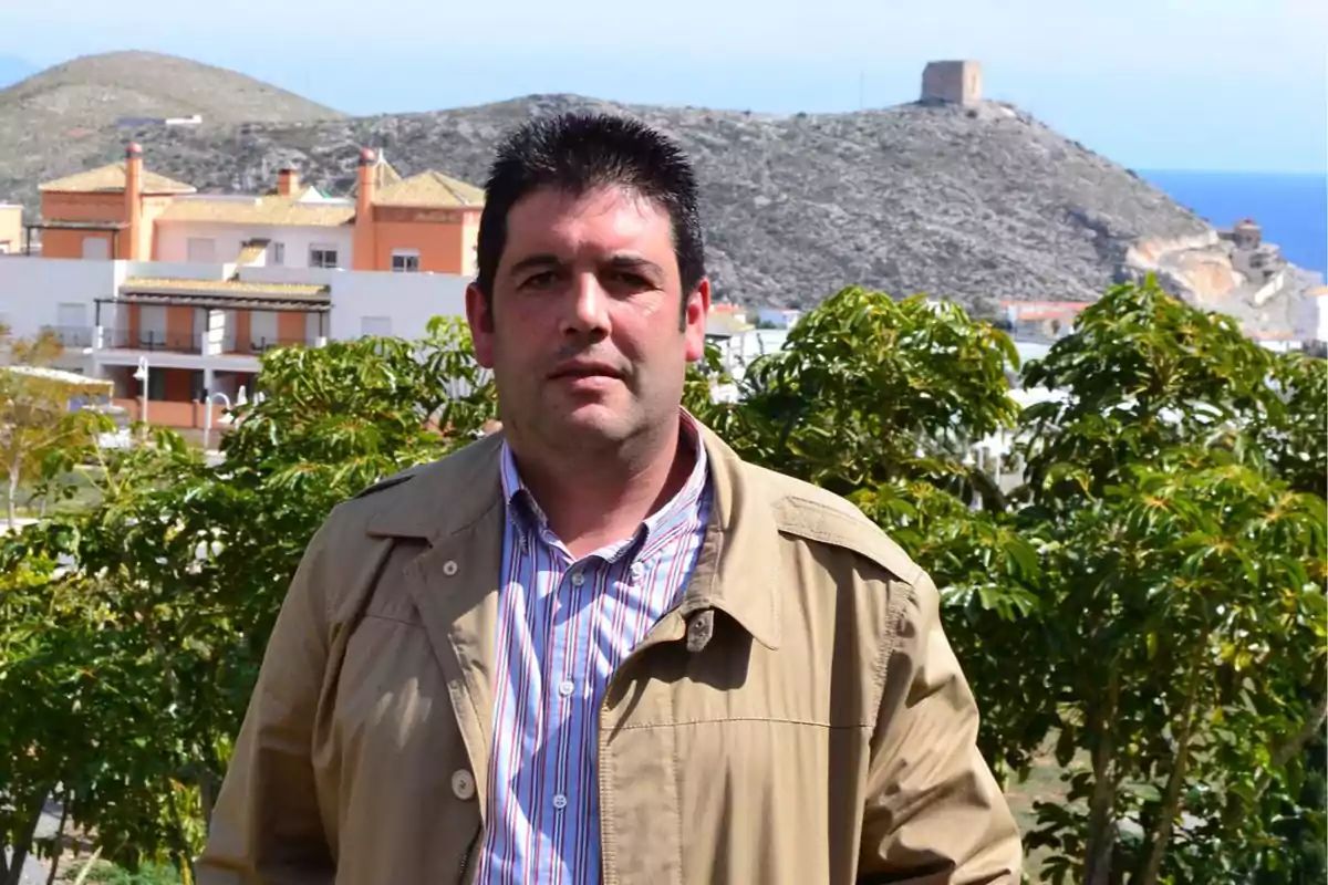 Hombre de cabello oscuro con chaqueta beige posando al aire libre con casas, vegetación y una colina al fondo