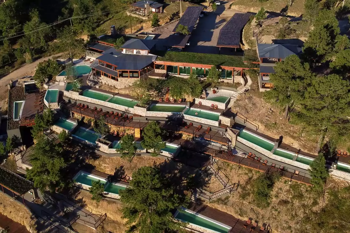 Vista aérea de un complejo turístico en la montaña con varias albercas en terrazas rodeadas de árboles y caminos de madera