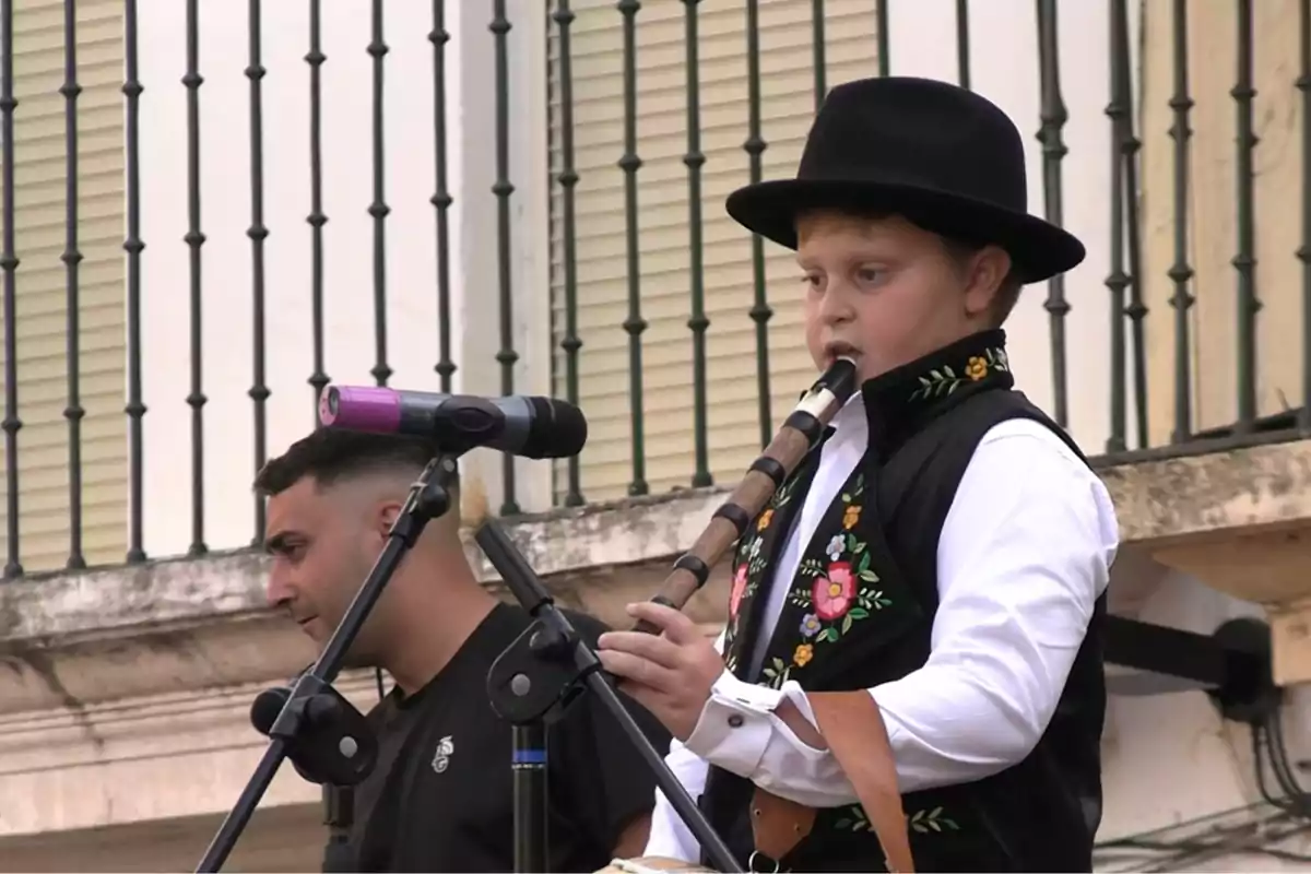 Niño con sombrero negro y chaleco bordado tocando una flauta tradicional frente a un micrófono en un escenario al aire libre Niño con sombrero negro y chaleco bordado tocando una flauta tradicional frente a un micrófono en un escenario al aire libre