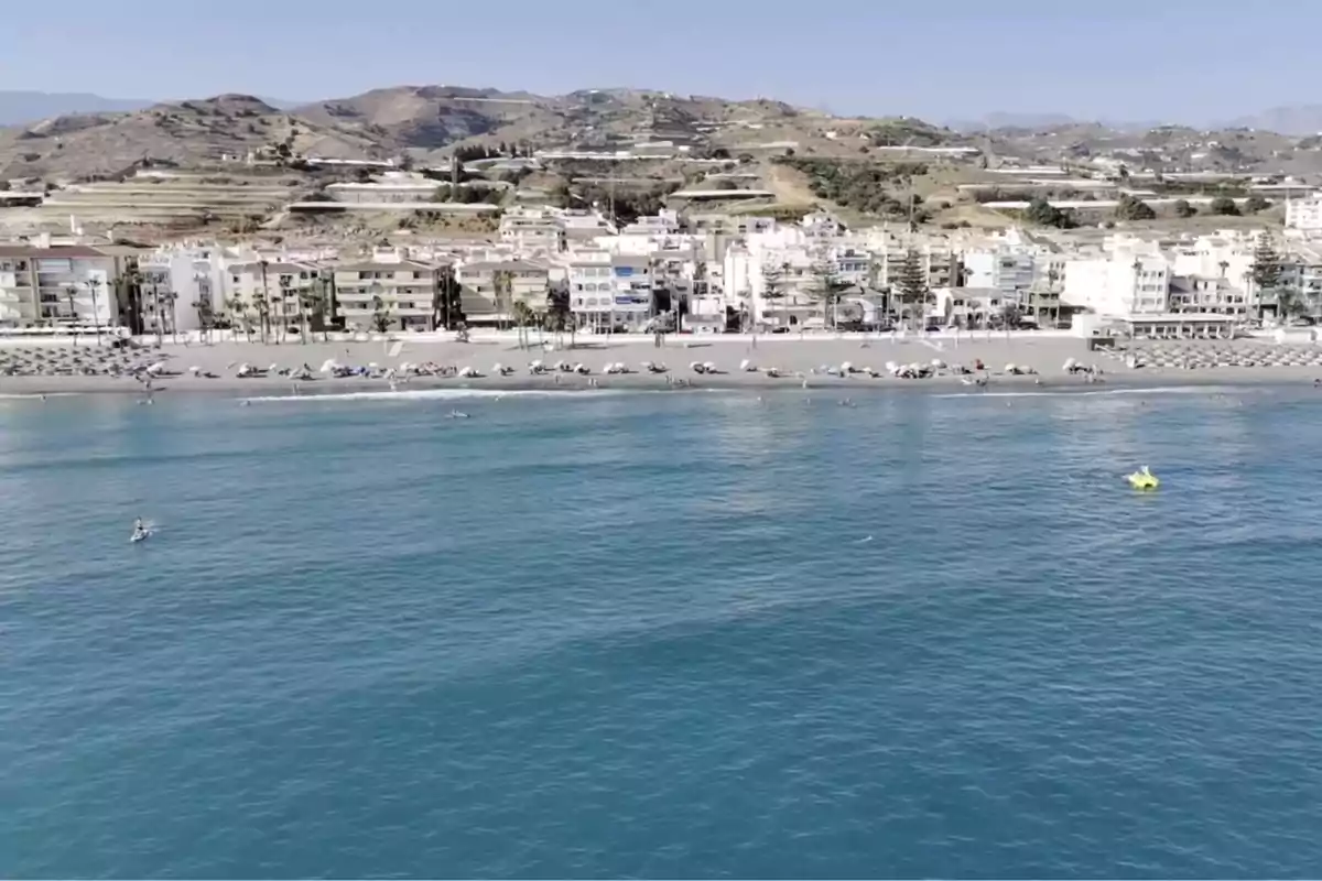 Vista de una playa con sombrillas y edificios al fondo, rodeada de colinas y mar azul en primer plano Vista de una playa con sombrillas y edificios al fondo, rodeada de colinas y mar azul en primer plano