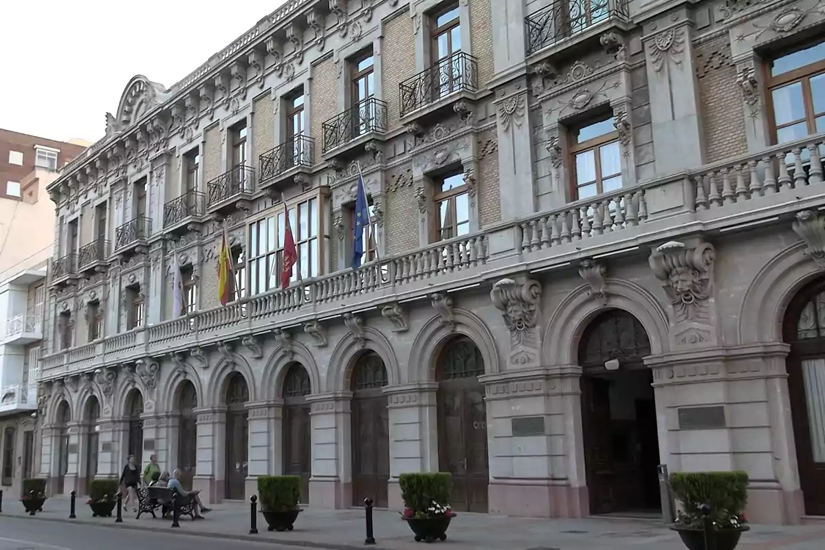 Fachada de un edificio histórico de varios pisos con balcones de hierro forjado, arcos en la planta baja, varias banderas y personas caminando por la acera. Fachada de un edificio histórico de varios pisos con balcones de hierro forjado, arcos en la planta baja, varias banderas y personas caminando por la acera.