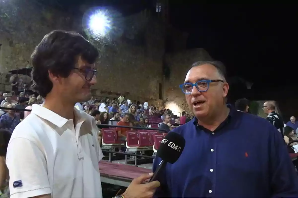 Un joven con gafas y camisa blanca entrevista a un hombre con gafas azules y camisa azul en un evento al aire libre con público sentado al fondo Un joven con gafas y camisa blanca entrevista a un hombre con gafas azules y camisa azul en un evento al aire libre con público sentado al fondo