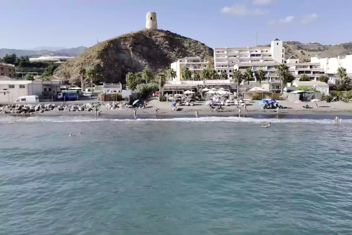 Playa con personas disfrutando del mar y edificios al fondo, en una colina se observa una torre antigua Playa con personas disfrutando del mar y edificios al fondo, en una colina se observa una torre antigua