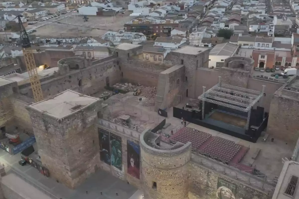 Vista aérea de un castillo antiguo con un escenario y asientos instalados en el patio interior, rodeado de murallas de piedra y con una grúa visible en una esquina, al fondo se observa una ciudad con casas y calles. Vista aérea de un castillo antiguo con un escenario y asientos instalados en el patio interior, rodeado de murallas de piedra y con una grúa visible en una esquina, al fondo se observa una ciudad con casas y calles.
