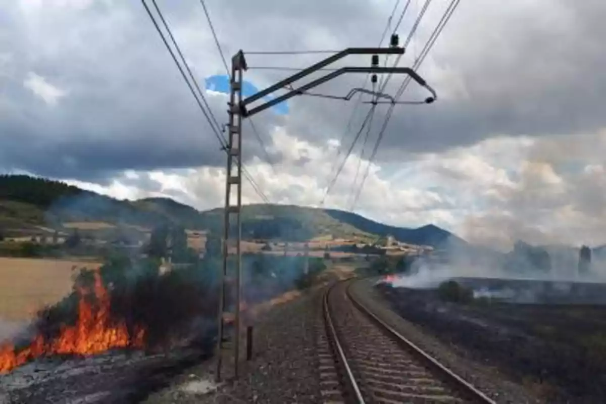 Incendio junto a unas vías de tren en una zona rural con colinas al fondo y cielo nublado Incendio junto a unas vías de tren en una zona rural con colinas al fondo y cielo nublado