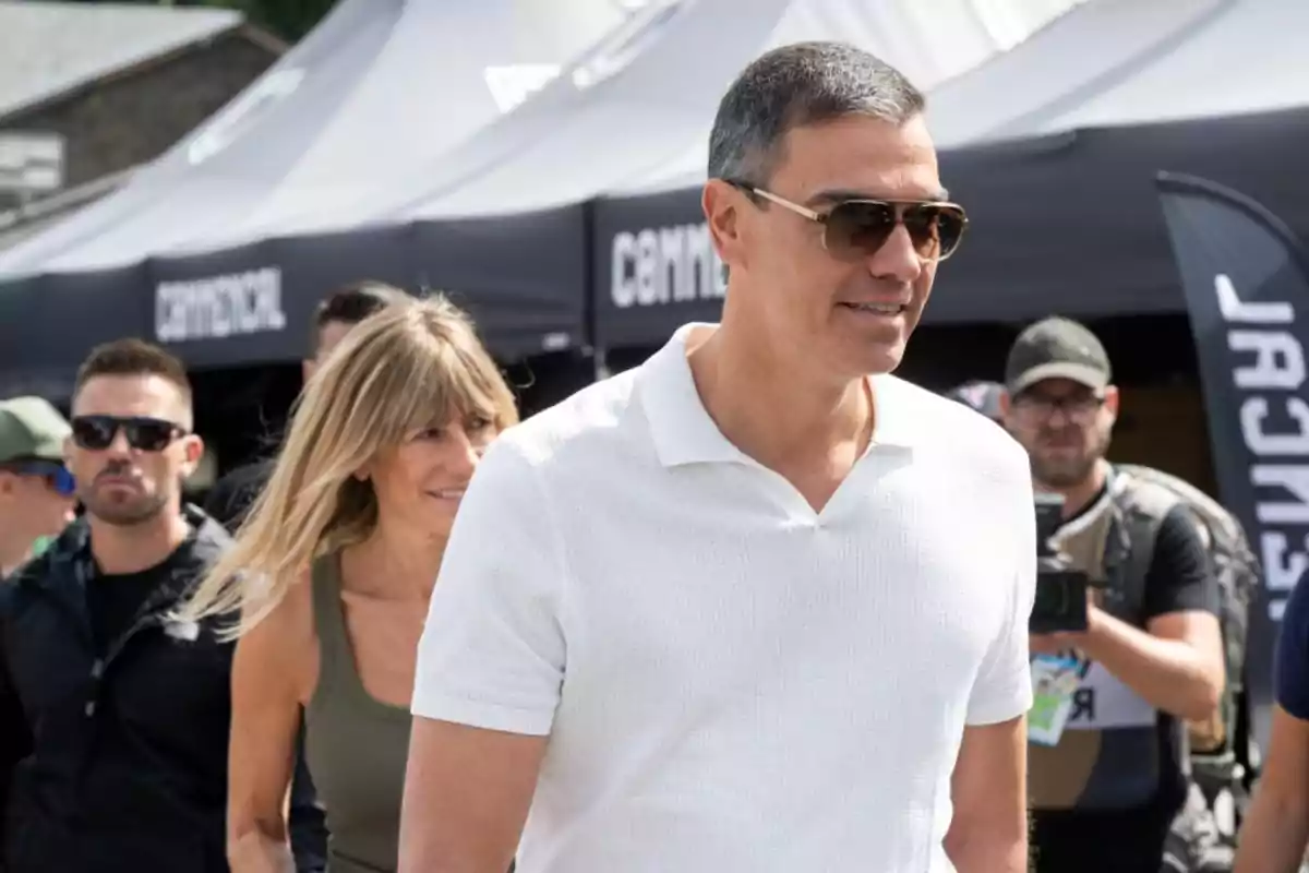Hombre con gafas de sol y camisa blanca caminando al aire libre acompañado de varias personas en un evento Hombre con gafas de sol y camisa blanca caminando al aire libre acompañado de varias personas en un evento
