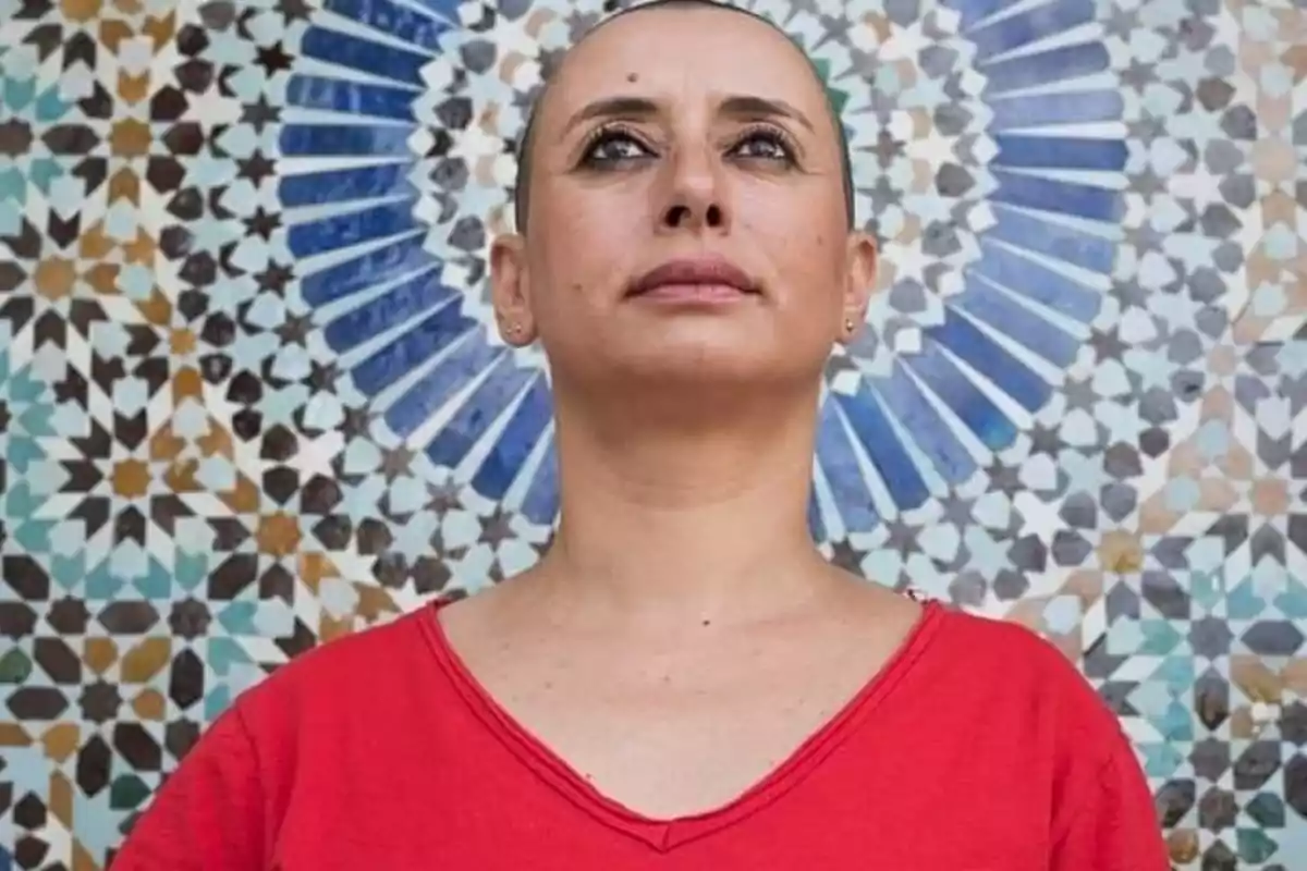 Mujer de cabello rapado con camiseta roja posando frente a un mosaico colorido de patrones geométricos Mujer de cabello rapado con camiseta roja posando frente a un mosaico colorido de patrones geométricos