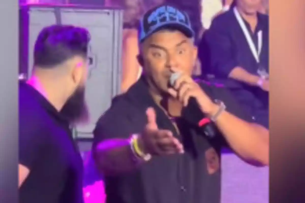Un cantante con gorra azul sostiene un micrófono mientras extiende la mano hacia el público en un escenario iluminado. Un cantante con gorra azul sostiene un micrófono mientras extiende la mano hacia el público en un escenario iluminado.