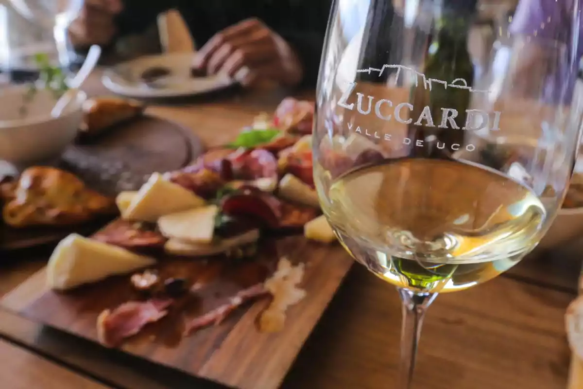 Copa de vino blanco con la marca Zuccardi grabada, al fondo una tabla de quesos y embutidos sobre una mesa de madera Copa de vino blanco con la marca Zuccardi grabada, al fondo una tabla de quesos y embutidos sobre una mesa de madera