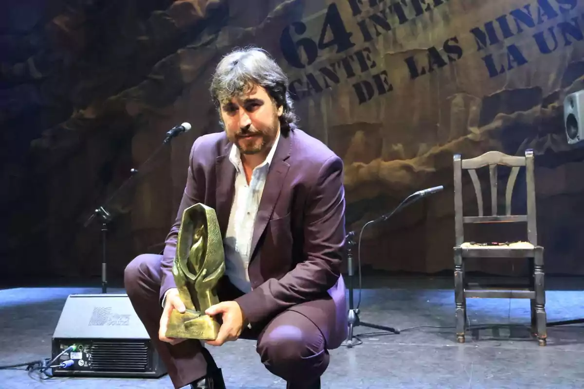 Hombre con traje sosteniendo un trofeo en el escenario de un festival de flamenco con una silla y un micrófono al fondo Hombre con traje sosteniendo un trofeo en el escenario de un festival de flamenco con una silla y un micrófono al fondo
