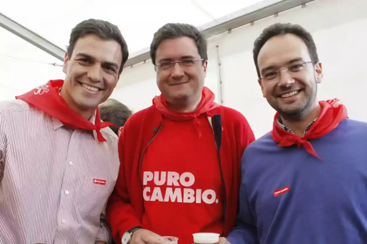 Tres hombres sonríen a la cámara usando pañuelos rojos en el cuello y ropa casual, uno de ellos lleva una sudadera roja con el texto PURO CAMBIO Tres hombres sonríen a la cámara usando pañuelos rojos en el cuello y ropa casual, uno de ellos lleva una sudadera roja con el texto PURO CAMBIO