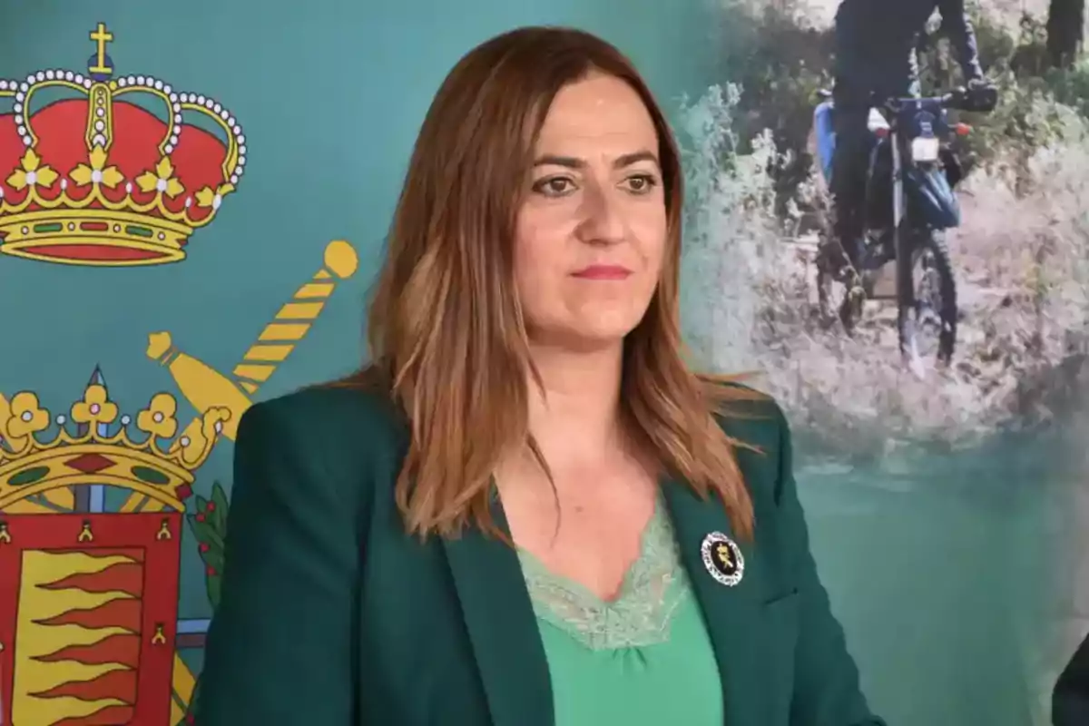 Mujer de cabello castaño y liso con chaqueta verde posando frente a un fondo institucional con un escudo y una imagen de una persona en motocicleta. Mujer de cabello castaño y liso con chaqueta verde posando frente a un fondo institucional con un escudo y una imagen de una persona en motocicleta.