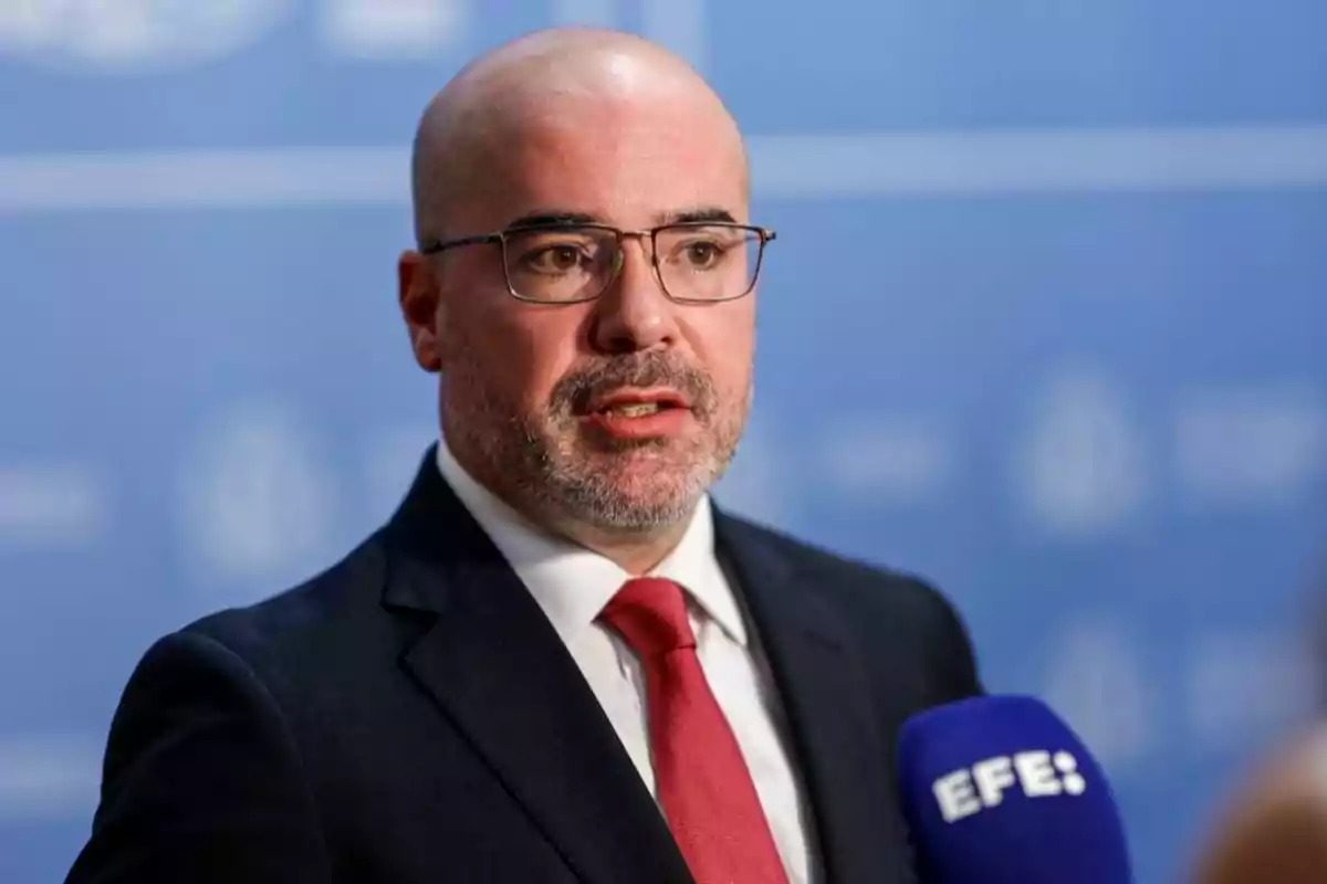 Hombre calvo con gafas y barba corta usando traje oscuro y corbata roja hablando frente a un micrófono azul con el logo de EFE sobre un fondo azul desenfocado Hombre calvo con gafas y barba corta usando traje oscuro y corbata roja hablando frente a un micrófono azul con el logo de EFE sobre un fondo azul desenfocado