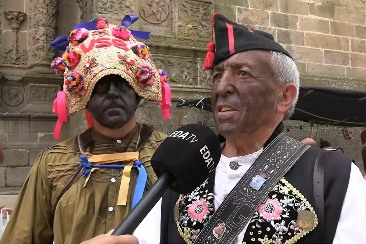 Dos hombres con la cara pintada de negro llevan trajes tradicionales y uno de ellos es entrevistado con un micrófono que tiene el logo de EDA TV mientras el otro lleva un sombrero decorado con flores de colores y cintas frente a una pared de piedra tallada. Dos hombres con la cara pintada de negro llevan trajes tradicionales y uno de ellos es entrevistado con un micrófono que tiene el logo de EDA TV mientras el otro lleva un sombrero decorado con flores de colores y cintas frente a una pared de piedra tallada.
