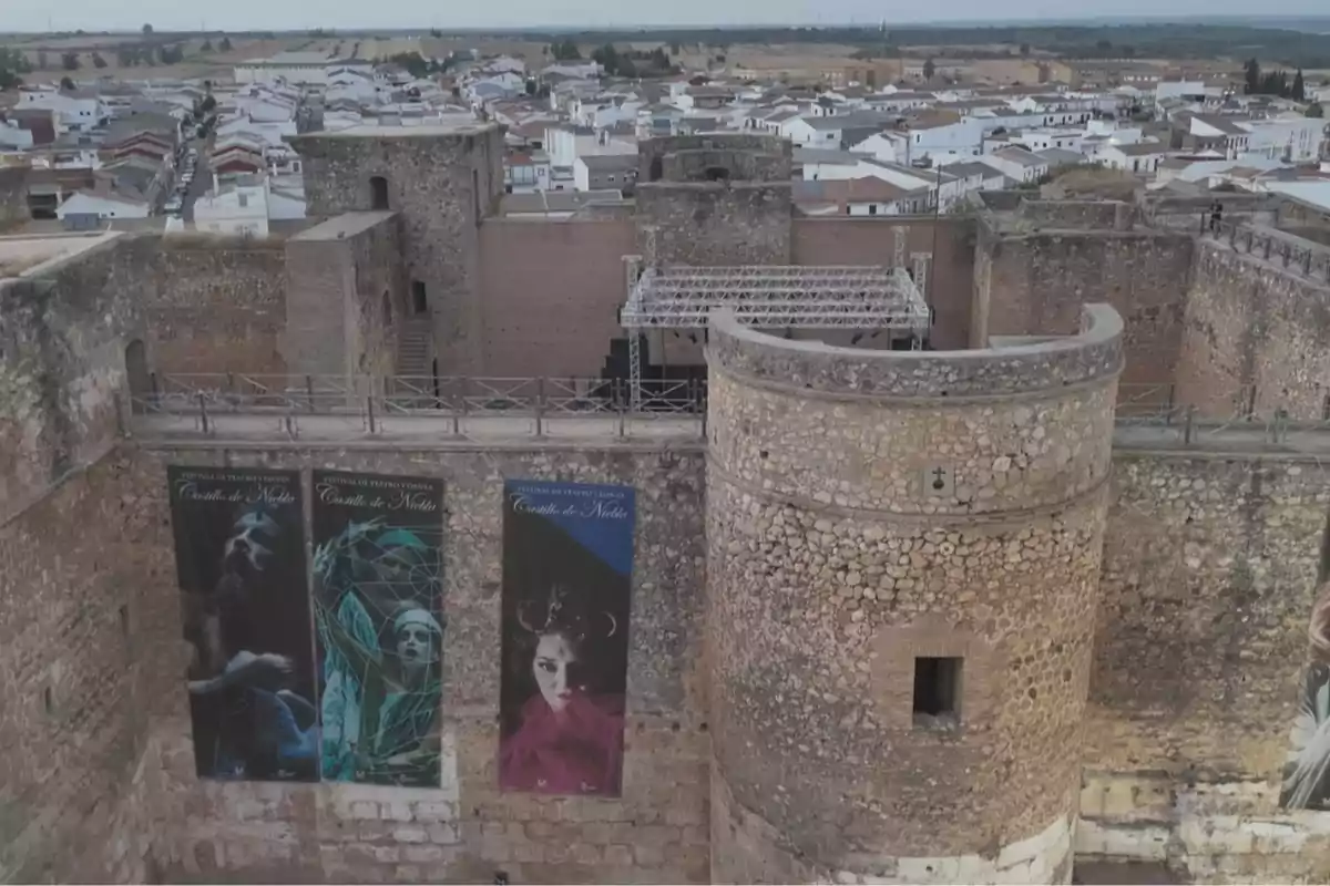 Vista aérea de un castillo de piedra con murallas y torres, en cuyo interior hay un escenario y tres grandes carteles de un festival, al fondo se observa un pueblo con casas blancas Vista aérea de un castillo de piedra con murallas y torres, en cuyo interior hay un escenario y tres grandes carteles de un festival, al fondo se observa un pueblo con casas blancas