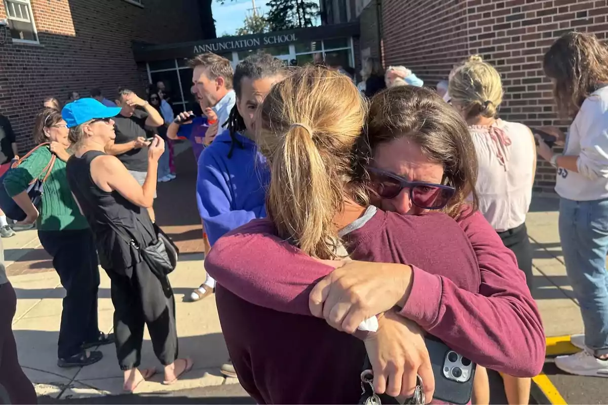 Varias personas se encuentran afuera de un edificio escolar, dos mujeres en primer plano se abrazan con fuerza y una de ellas parece estar llorando, mientras otras personas conversan o esperan alrededor. Varias personas se encuentran afuera de un edificio escolar, dos mujeres en primer plano se abrazan con fuerza y una de ellas parece estar llorando, mientras otras personas conversan o esperan alrededor.