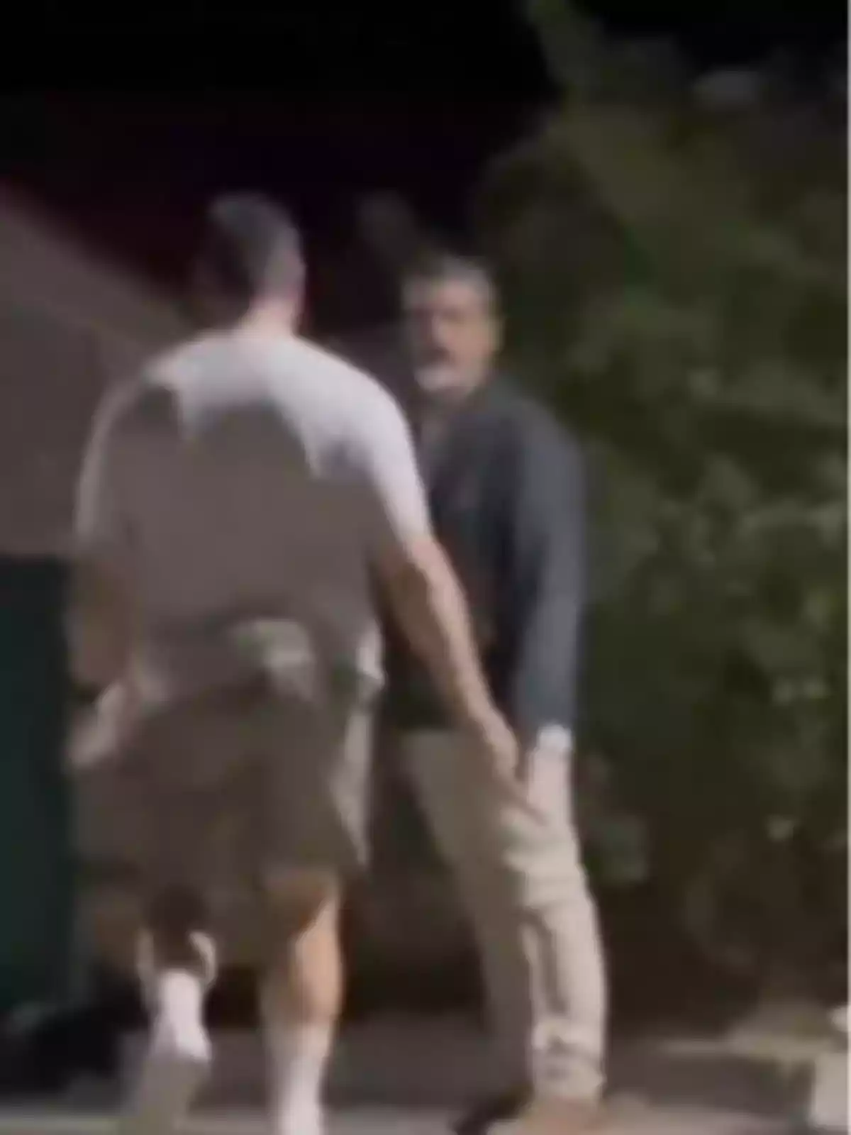 Dos hombres de pie conversan al aire libre durante la noche, uno de ellos lleva camiseta clara y pantalones cortos, el otro viste ropa oscura y pantalón largo, la imagen está desenfocada. Dos hombres de pie conversan al aire libre durante la noche, uno de ellos lleva camiseta clara y pantalones cortos, el otro viste ropa oscura y pantalón largo, la imagen está desenfocada.