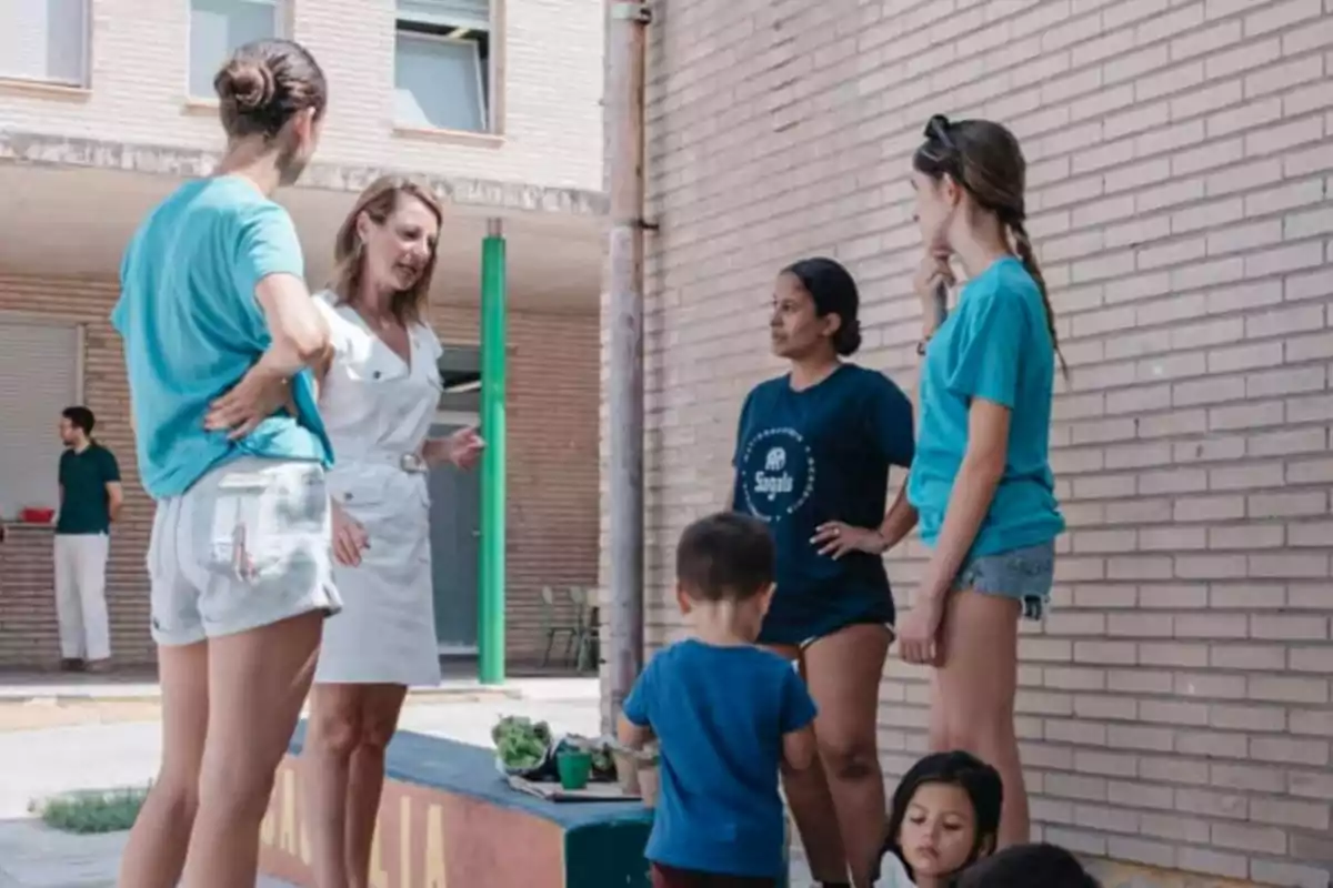 Varias personas conversan y juegan con niños en un patio exterior junto a un edificio de ladrillo