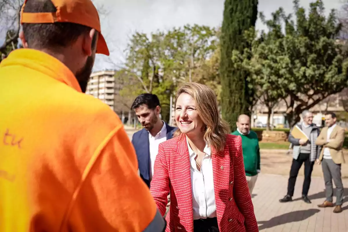 Una mujer sonriente con chaqueta roja saluda a un hombre con ropa naranja en un parque mientras otras personas observan al fondo