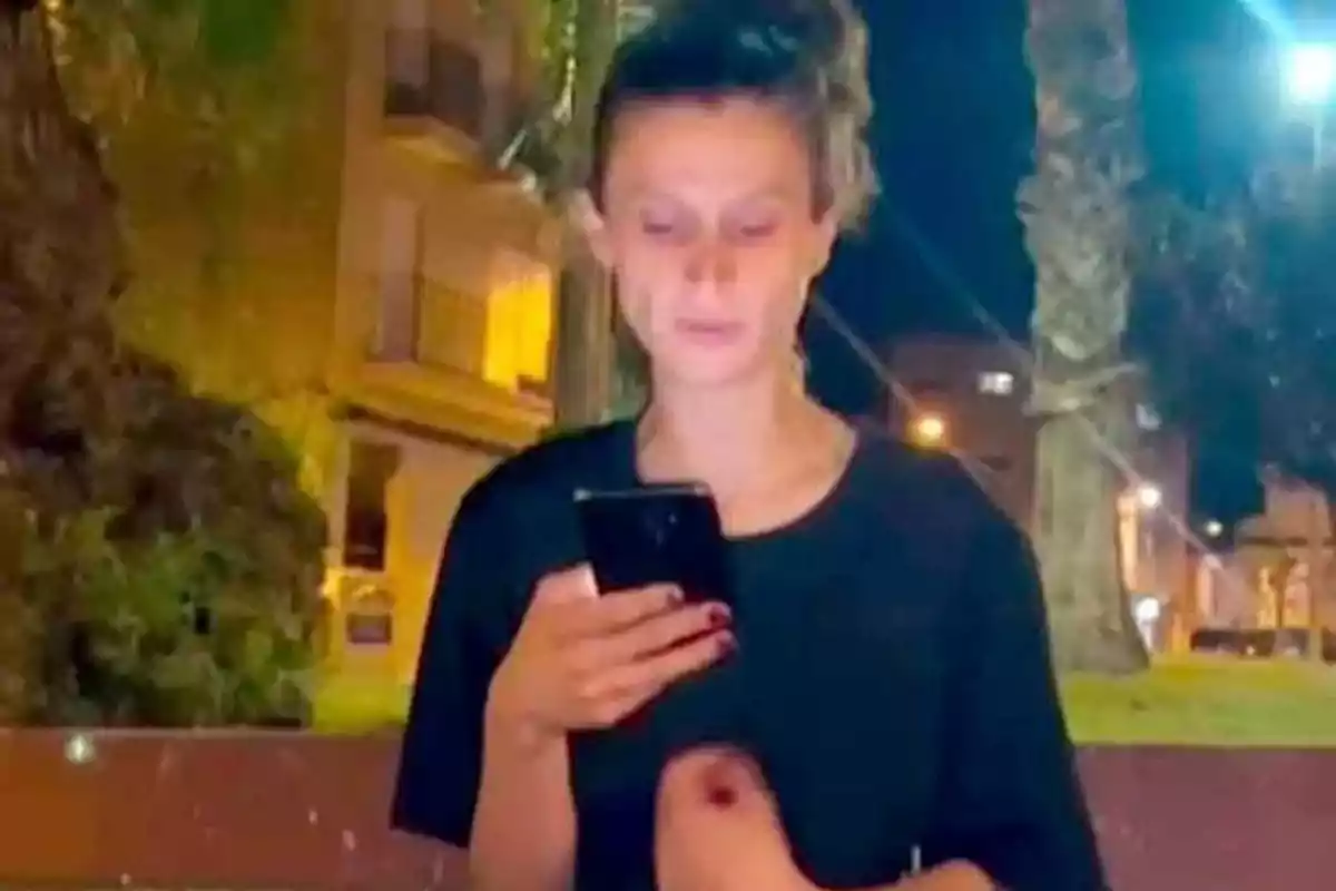 Mujer joven de pie al aire libre en la noche mirando su teléfono móvil con edificios y árboles al fondo Mujer joven de pie al aire libre en la noche mirando su teléfono móvil con edificios y árboles al fondo