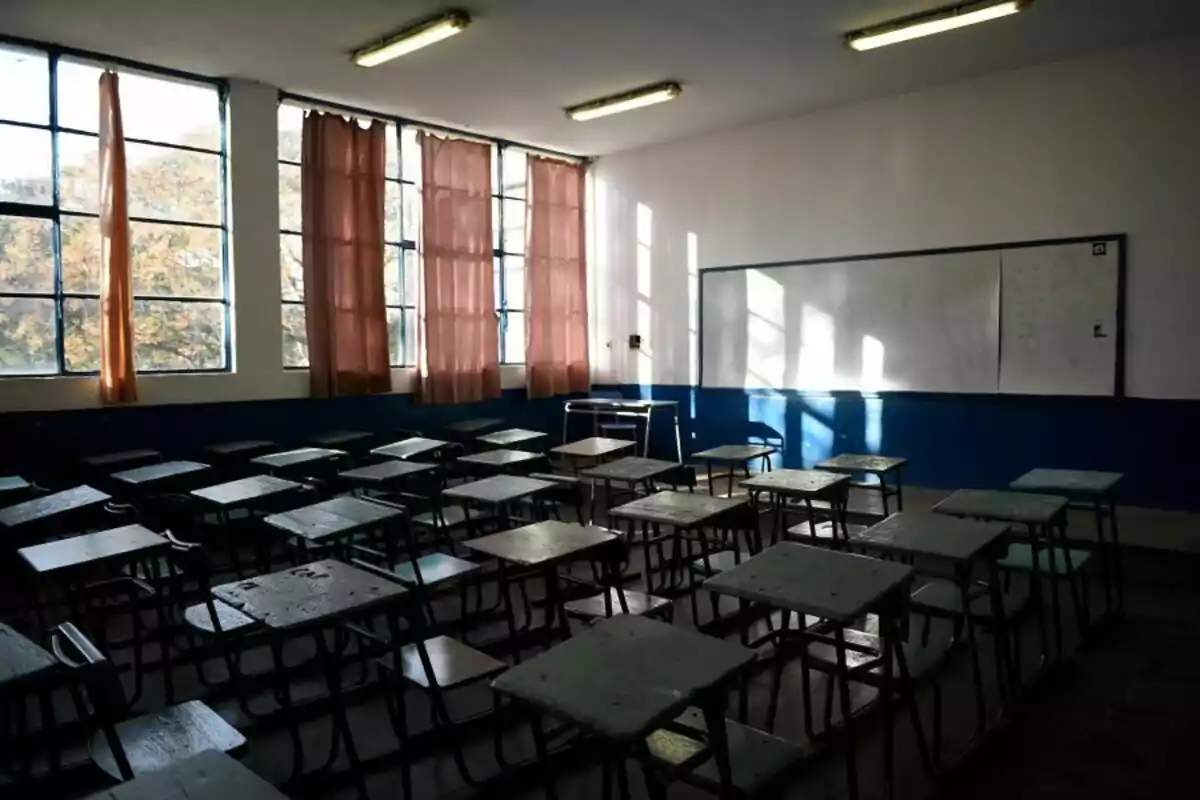 Aula vacía con escritorios y sillas desordenados, ventanas grandes con cortinas rosadas y un pizarrón blanco al fondo Aula vacía con escritorios y sillas desordenados, ventanas grandes con cortinas rosadas y un pizarrón blanco al fondo