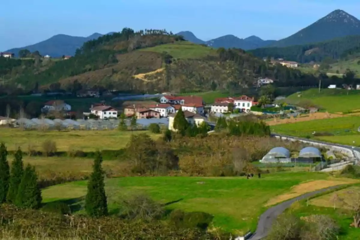 Vista de un pequeño pueblo rodeado de colinas verdes y montañas al fondo con casas de tejados rojos y campos de cultivo Vista de un pequeño pueblo rodeado de colinas verdes y montañas al fondo con casas de tejados rojos y campos de cultivo