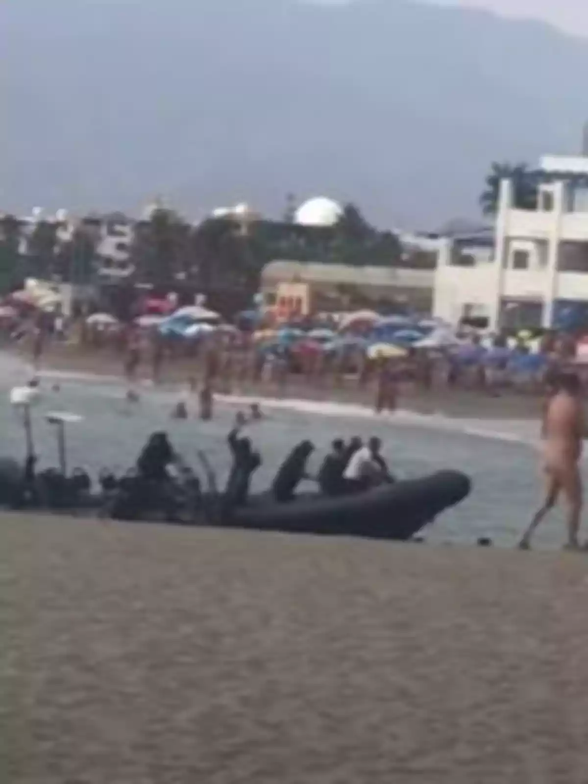 Un grupo de personas vestidas de negro llega a la orilla de una playa en una lancha inflable mientras varias personas disfrutan del mar y la arena en el fondo Un grupo de personas vestidas de negro llega a la orilla de una playa en una lancha inflable mientras varias personas disfrutan del mar y la arena en el fondo