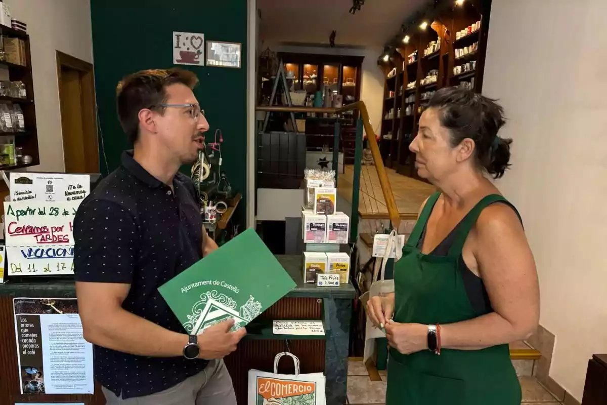 Un hombre con gafas sostiene una carpeta verde mientras conversa con una mujer que lleva un delantal verde dentro de una tienda con estantes llenos de productos y carteles informativos.