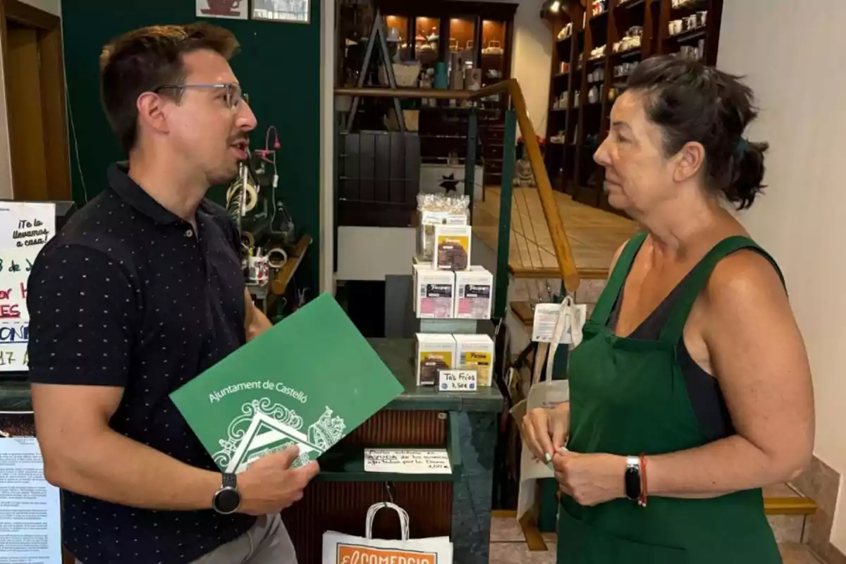 Un hombre con gafas sostiene una carpeta verde mientras conversa con una mujer vestida con delantal verde en el interior de una tienda Un hombre con gafas sostiene una carpeta verde mientras conversa con una mujer vestida con delantal verde en el interior de una tienda