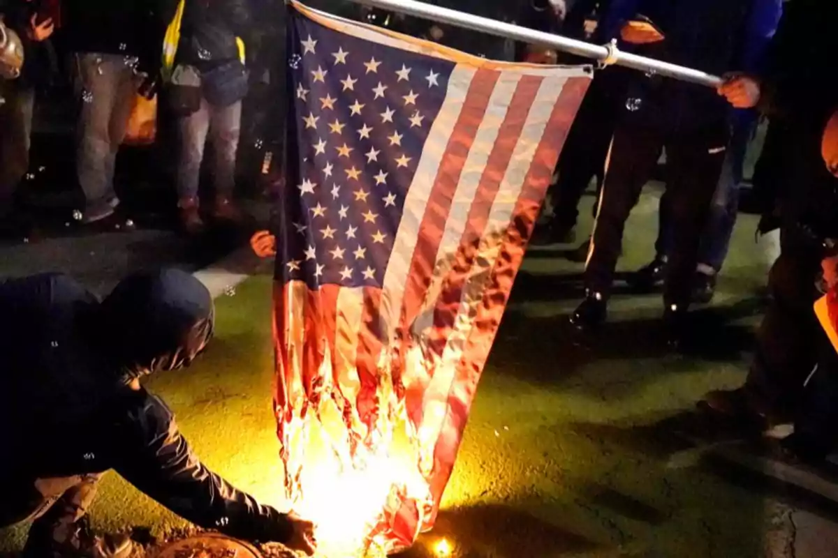 Varias personas rodean una bandera de Estados Unidos que está siendo quemada en el suelo durante una protesta nocturna Varias personas rodean una bandera de Estados Unidos que está siendo quemada en el suelo durante una protesta nocturna