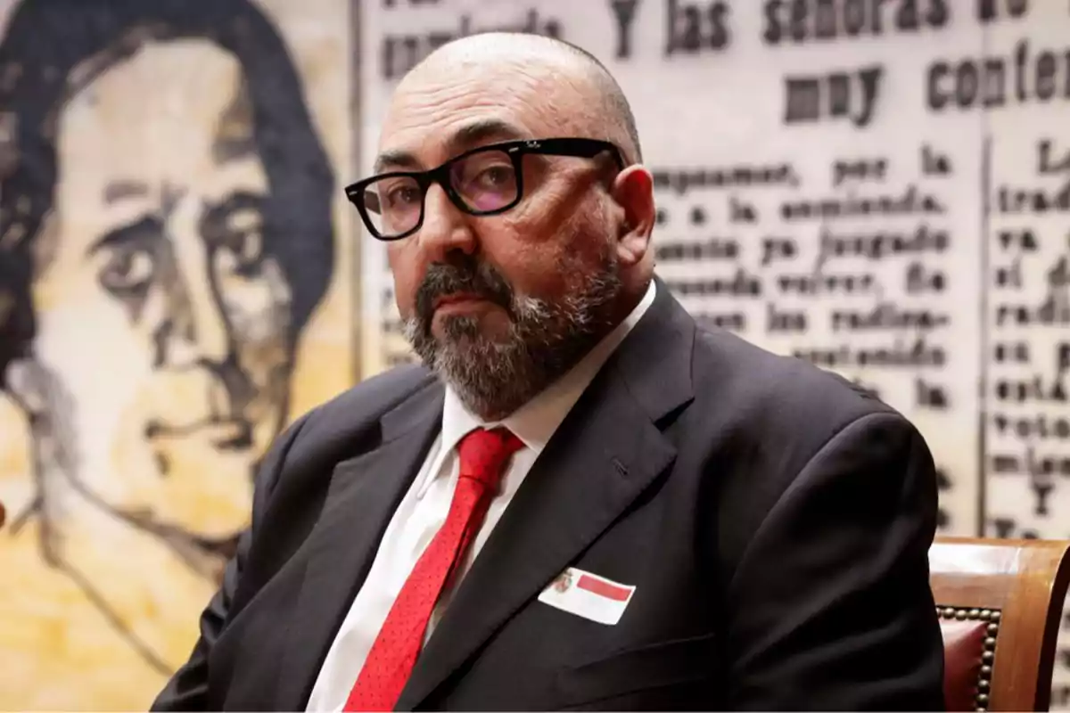 Hombre de barba y gafas con traje oscuro y corbata roja sentado frente a un fondo con texto y un retrato en blanco y negro.