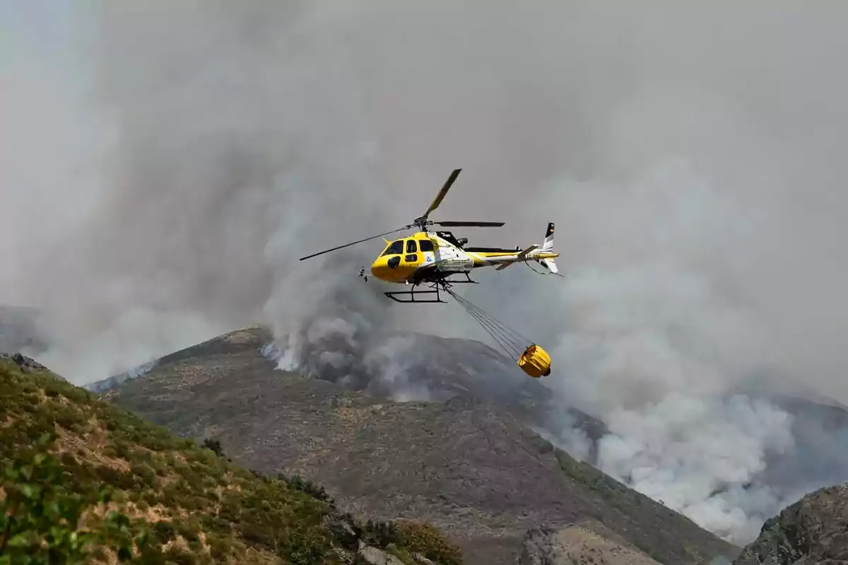 Helicóptero amarillo y blanco transportando un contenedor de agua sobre una zona montañosa afectada por un incendio forestal con humo denso en el fondo