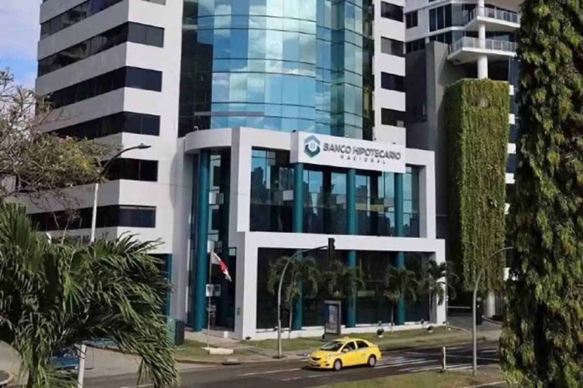 Fachada de un edificio moderno de vidrio y concreto con el letrero Banco Hipotecario Nacional y un taxi amarillo pasando por la calle frente al edificio