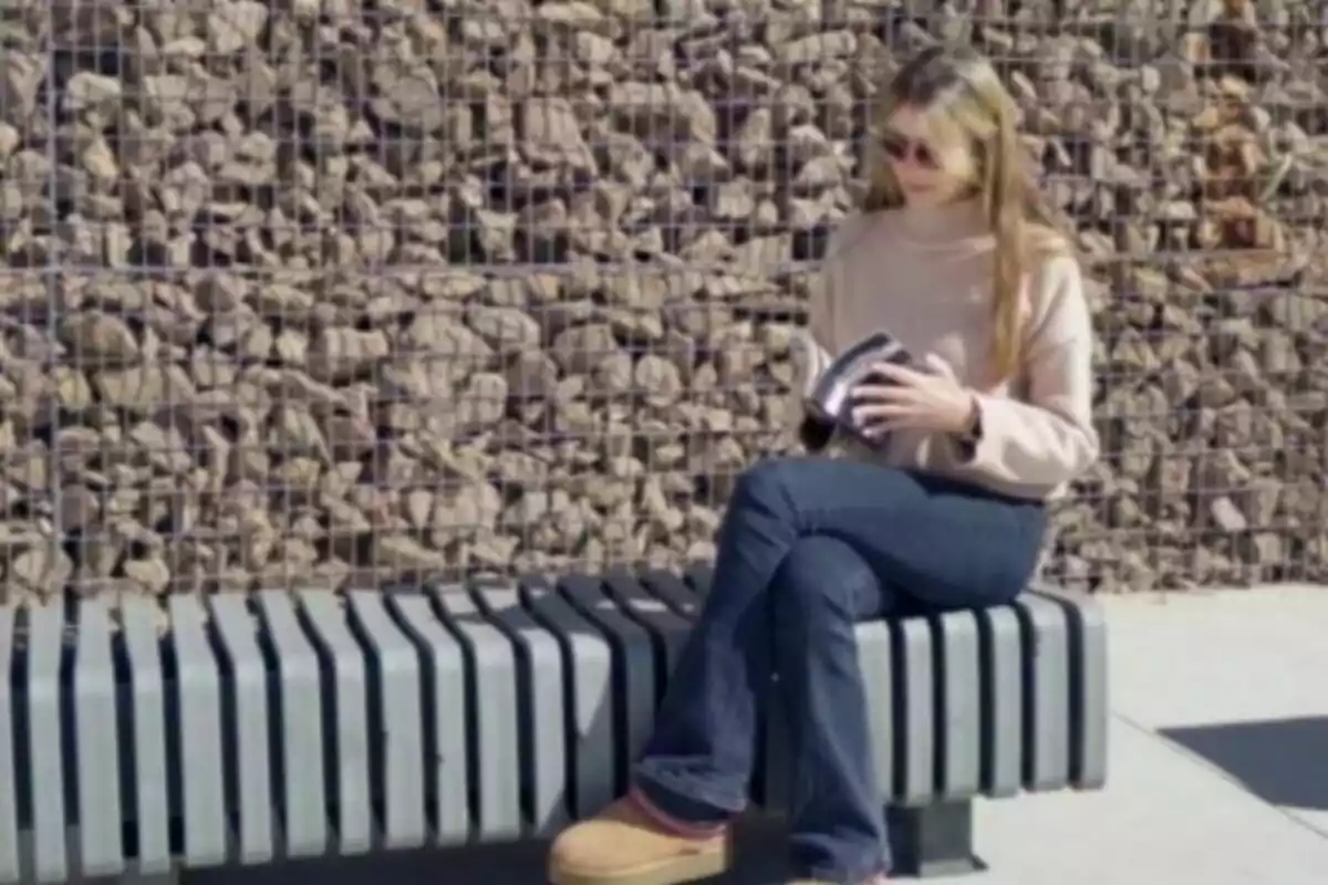 Mujer sentada en una banca al aire libre leyendo un libro frente a una pared de piedras Mujer sentada en una banca al aire libre leyendo un libro frente a una pared de piedras