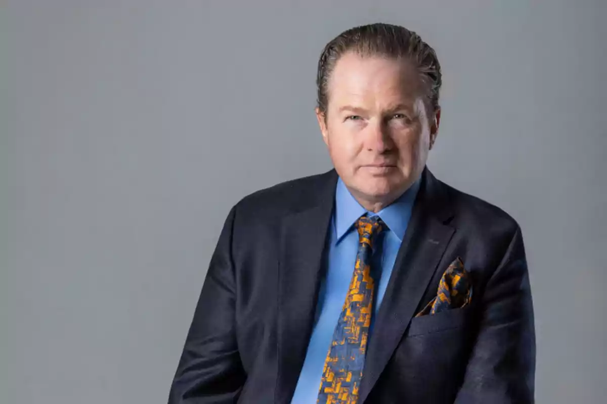 Hombre de mediana edad con cabello corto y peinado hacia atrás, usando traje azul oscuro, camisa azul y corbata con diseño naranja y azul, posando frente a un fondo gris.