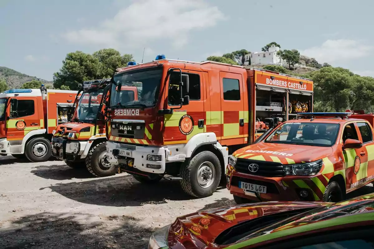 Varios vehículos de bomberos de color rojo y amarillo estacionados al aire libre junto a una zona arbolada y una casa blanca en una colina al fondo Varios vehículos de bomberos de color rojo y amarillo estacionados al aire libre junto a una zona arbolada y una casa blanca en una colina al fondo
