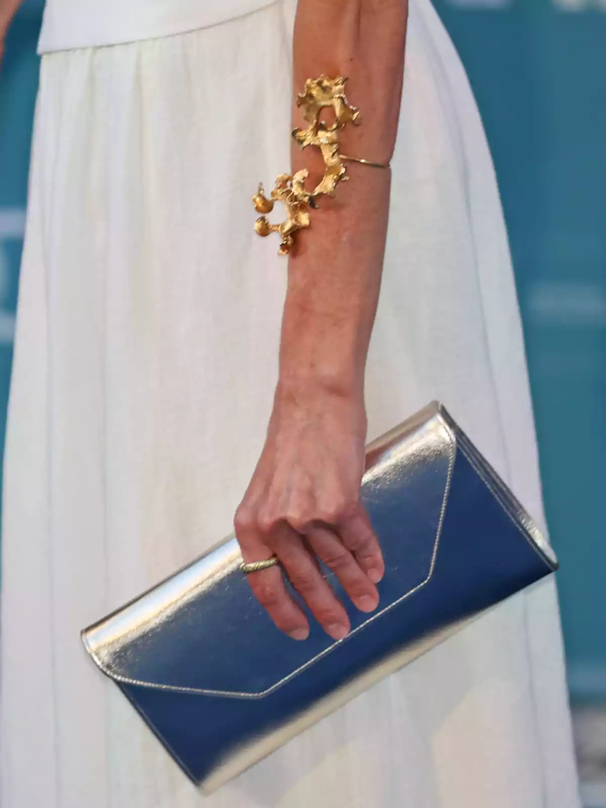 Queen Letizia in a white dress holding a metallic silver clutch and wearing a striking gold bracelet on her arm