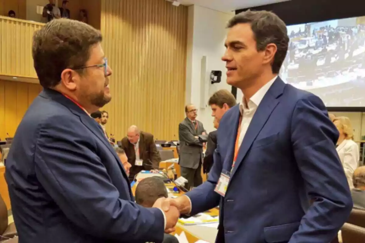 Dos hombres vestidos con traje se dan la mano en una sala de conferencias llena de personas Dos hombres vestidos con traje se dan la mano en una sala de conferencias llena de personas