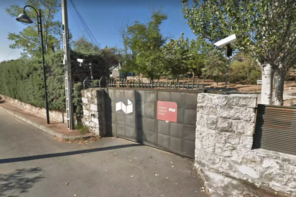 Portón metálico gris con letreros, rodeado de un muro de piedra y vegetación, cámaras de seguridad y un poste de luz en la acera Portón metálico gris con letreros, rodeado de un muro de piedra y vegetación, cámaras de seguridad y un poste de luz en la acera