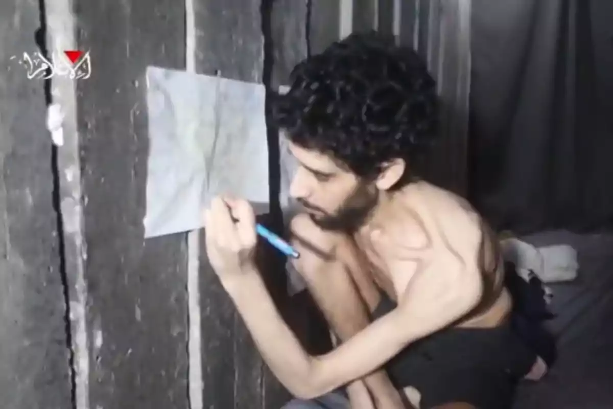 Hombre delgado y descalzo sentado en el suelo de una habitación oscura escribiendo en una hoja de papel pegada a la pared Hombre delgado y descalzo sentado en el suelo de una habitación oscura escribiendo en una hoja de papel pegada a la pared